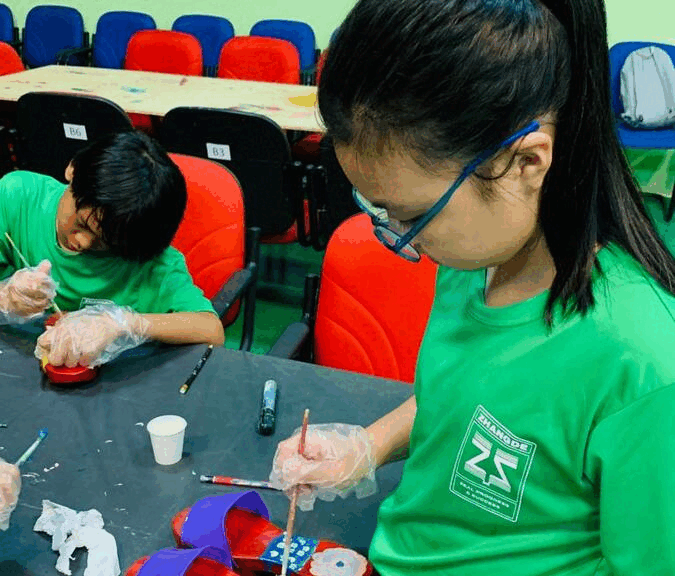 Two children in green shirts and plastic gloves painting crafts at a table in a colorful classroom.